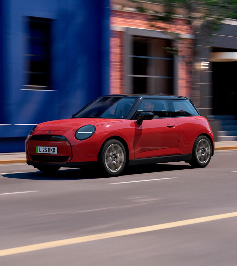 MINI Cooper electric hatchback driving on a city street with blue and brick buildings in the background, featuring black roof, circular LED headlights, and sleek modern design.